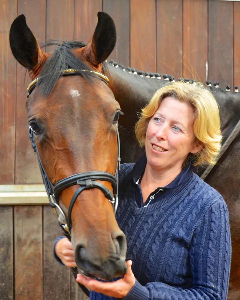 Alison Haste, smiling, holding the muzzle of a bay horse at a wooden stable